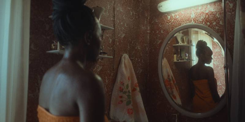 A Black woman is viewed from behind, her shoulders exposed. Her reflection is visible in the mirror before her.