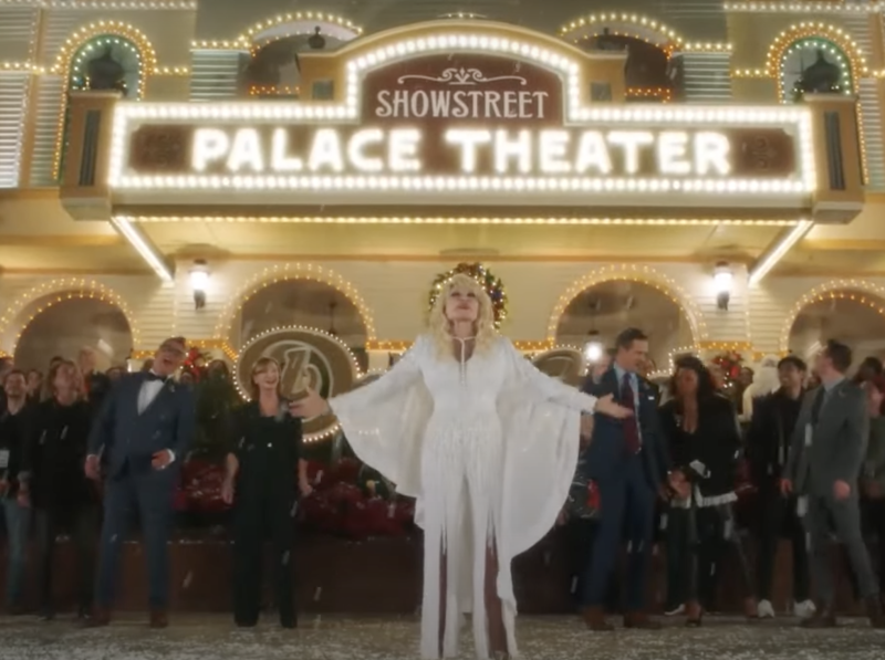 Dolly Parton stands under the marquee of the Palace Theater at Dollywood, surrounded by audiences members.