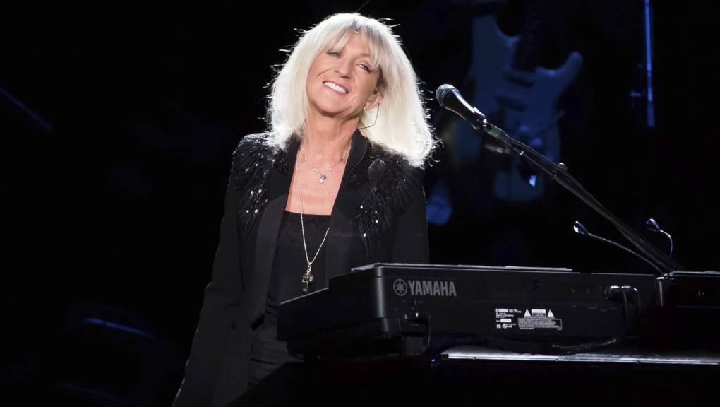a white woman with light blonde hair in a black sparkly outfit smiles at a piano onstage