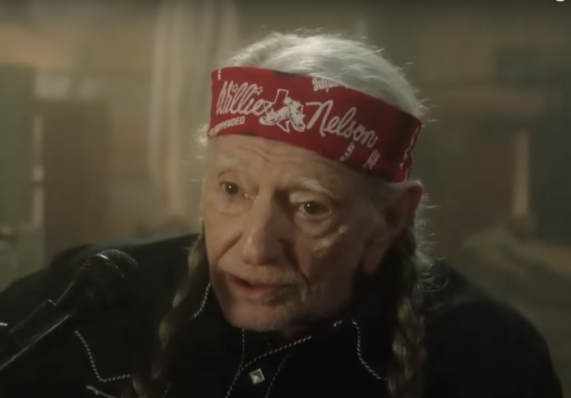 A senior man with long gray hair braided over his shoulders, wearing a red headband with the words Willie Nelson on it.