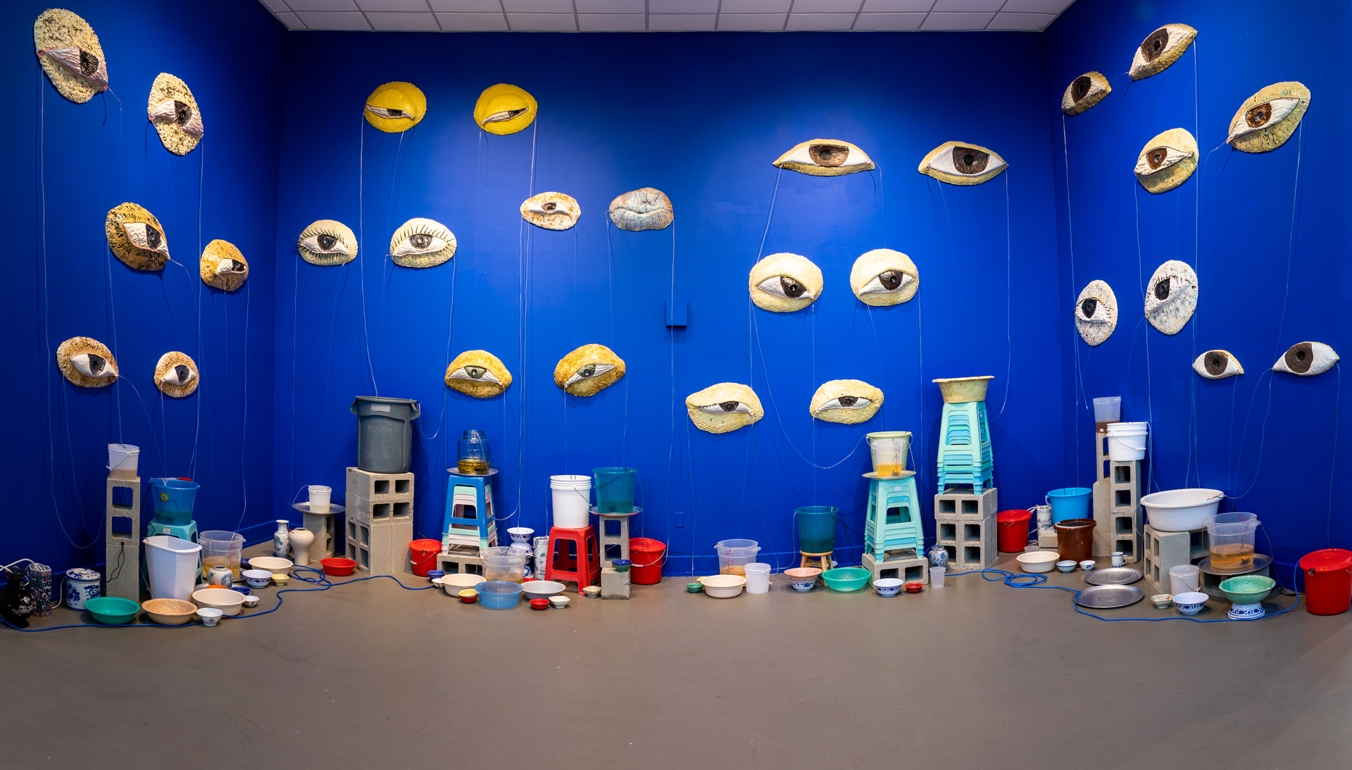 Blue gallery walls with large ceramic eyes mounted and multicolored buckets and stools below