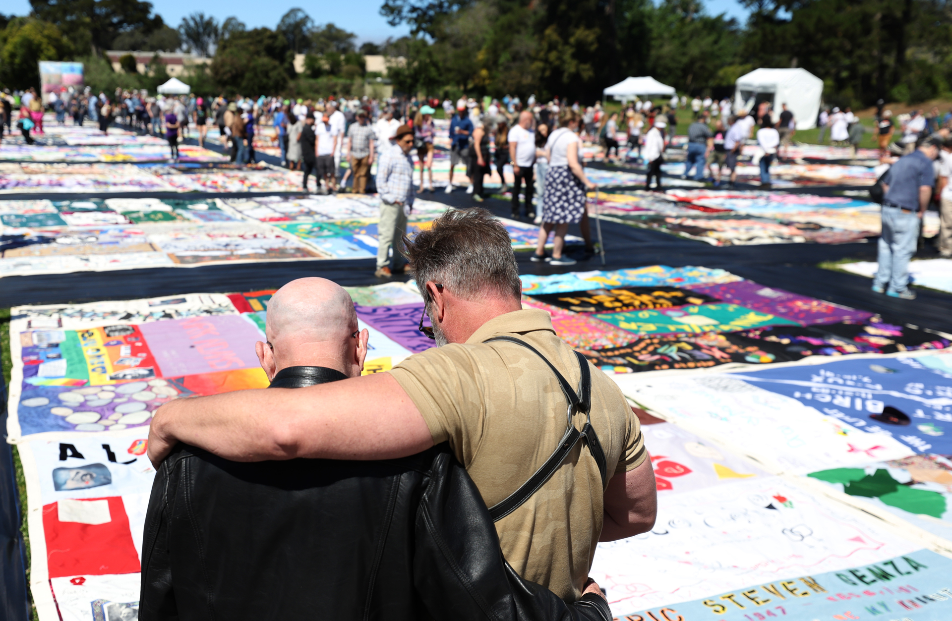 Two white men stand arm in arm looking down at embellished fabric panels, crowd in distance