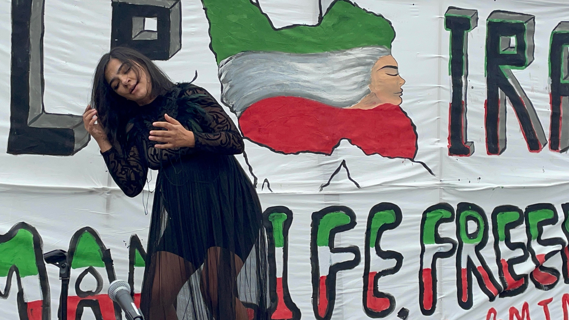 Visibly pregnant woman in black dress dances in front of mural depicting Iranian flag and protest messages