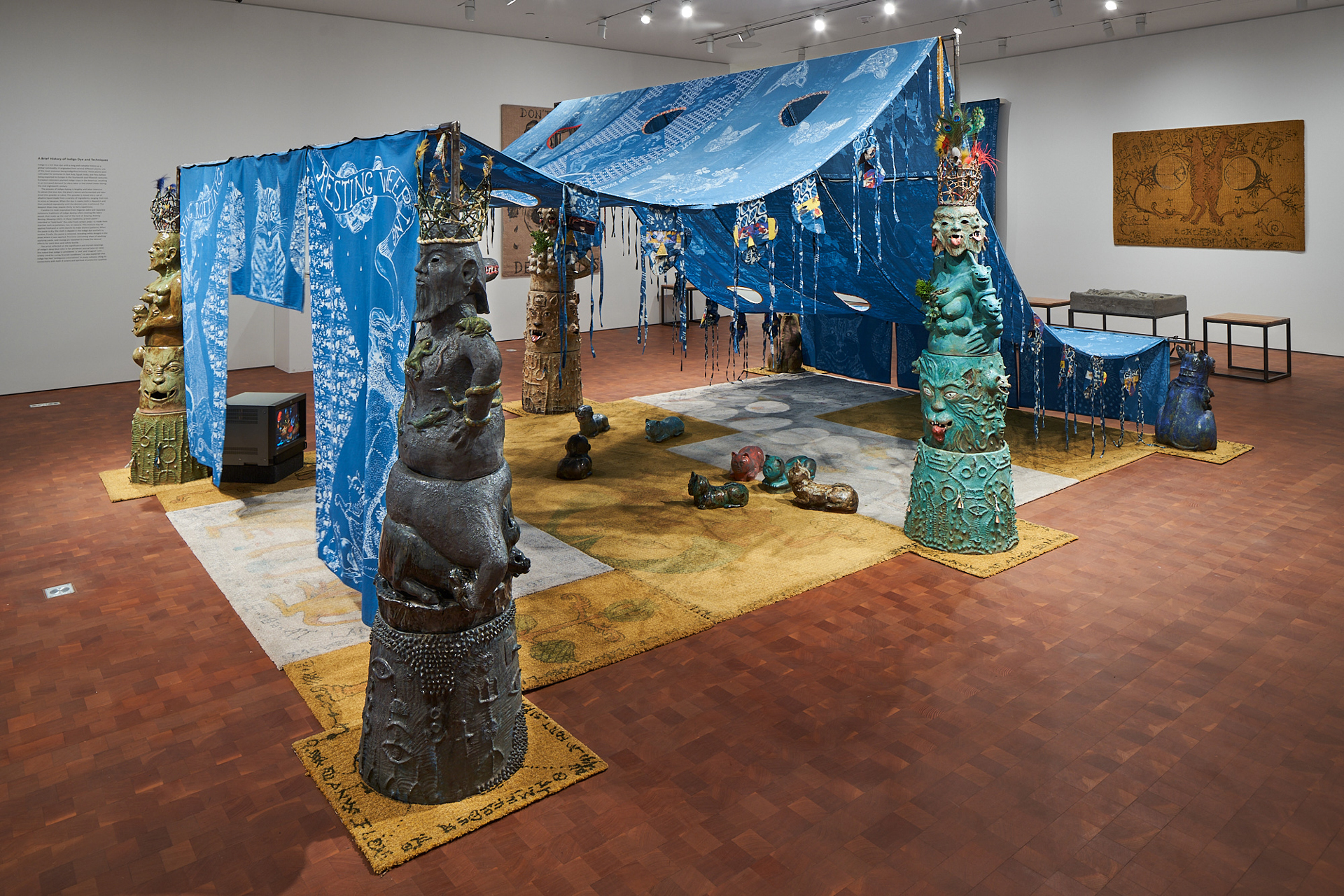 View of gallery with blue tent, large ceramic figures, a carpet and TV beneath and small audience of ceramic cats