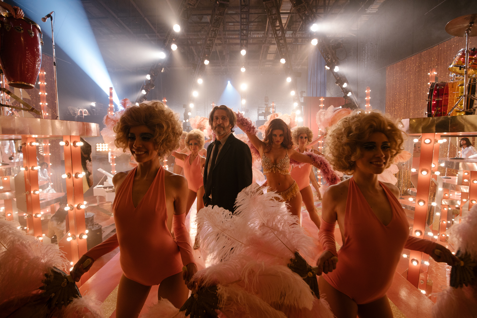 Older man in suit at center of dancers wearing pink leotards and feathers