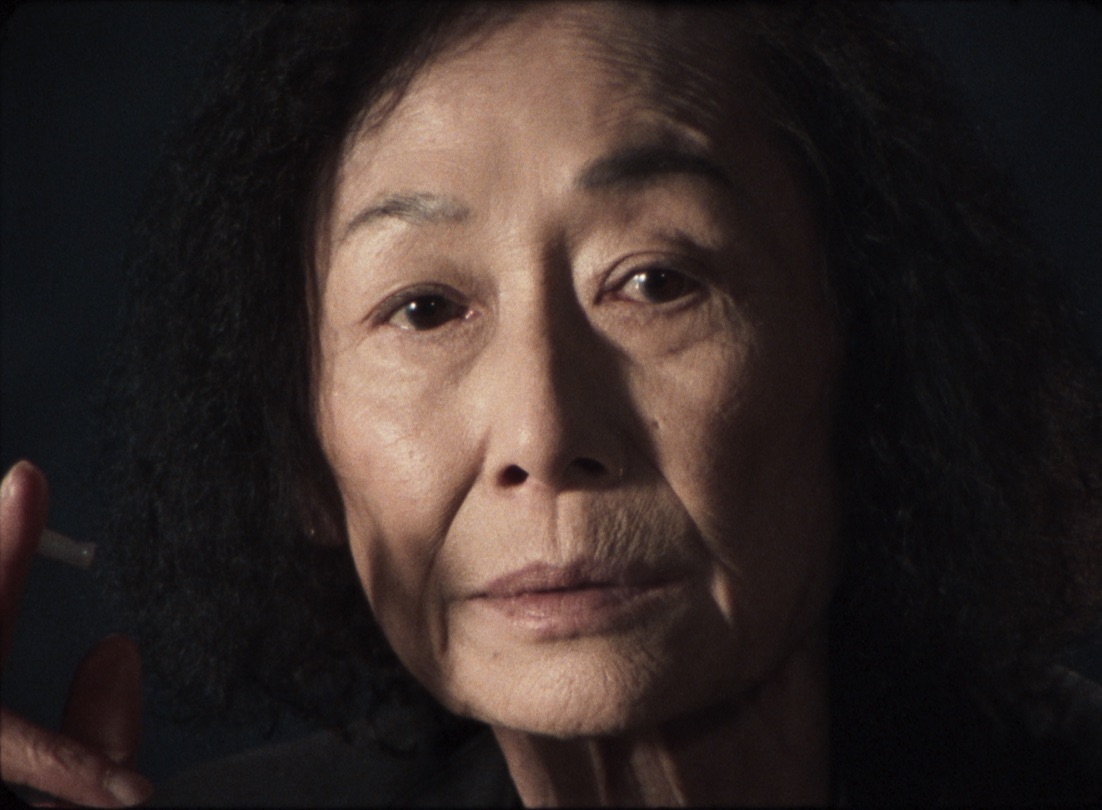 Older Asian woman holding a ciragrette, looking directly at camera with only face lit
