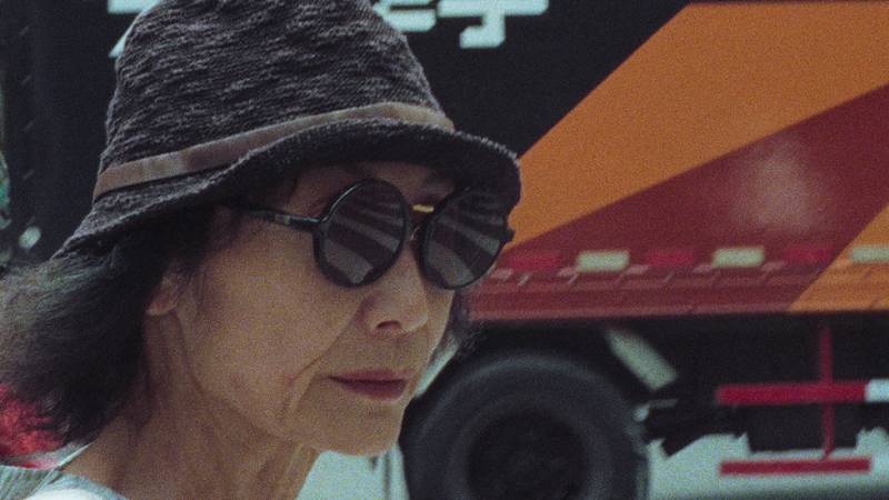 Grainy image of older Asian woman in hat and sunglasses with semitruck behind her