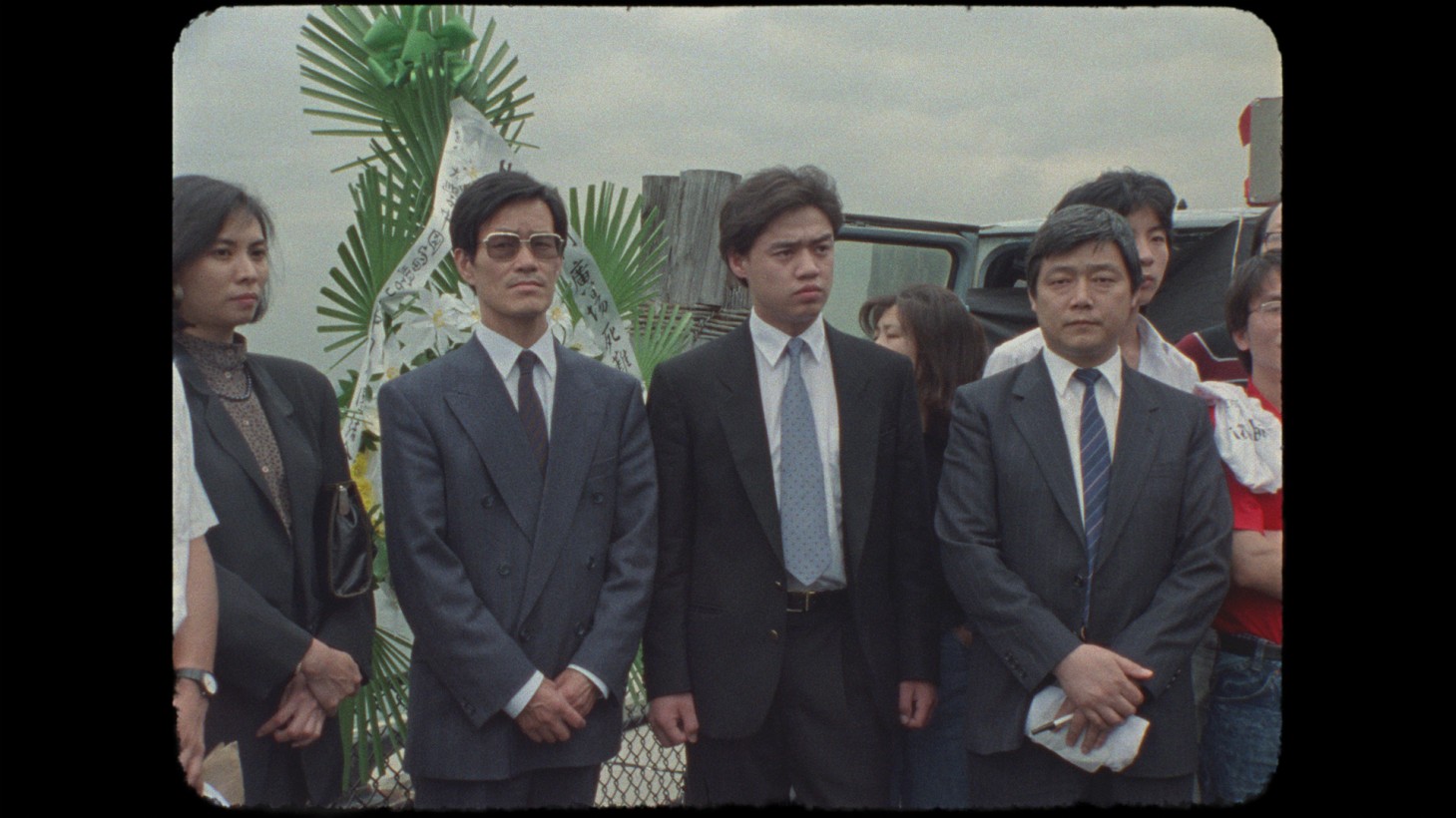 A group of unsmiling Asian men and women in suits, image from film cell with rounded corners