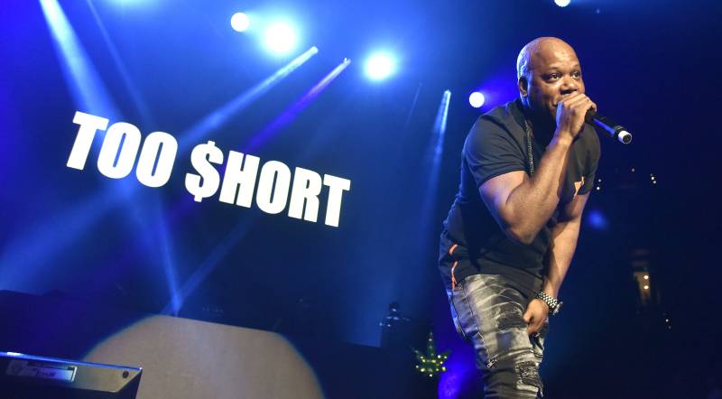 a Black man raps into a microphone on a stage with the words 'Too $hort' behind him in blue lights