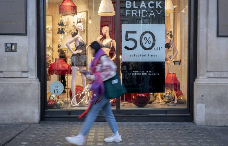 a person wearing jeans and white sneakers walks on a sidewalk in a city by a storefront with a sign that says 'black Friday 50% off'