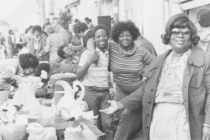Three African-American family members laughing and gesturing at items on a table during a flea market