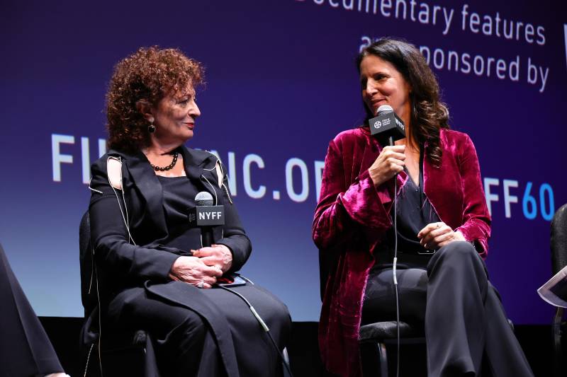 Two women holding microphones sits on basic chairs on stage, looking admiringly at one another.