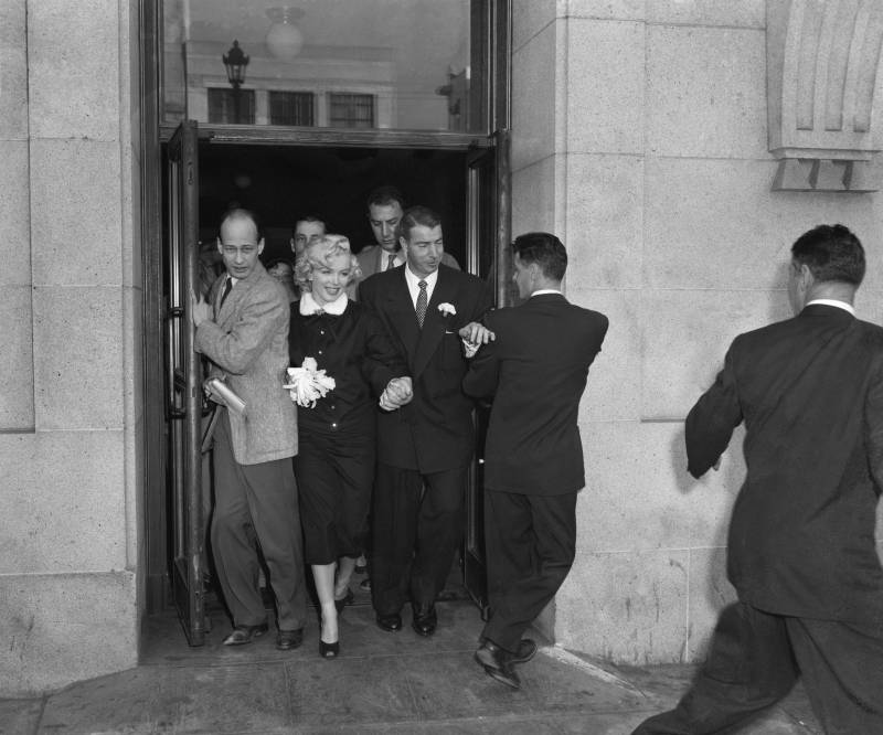 A couple dressed in smart 1950s clothing squeeze through a large door, surrounded by photographers and press.