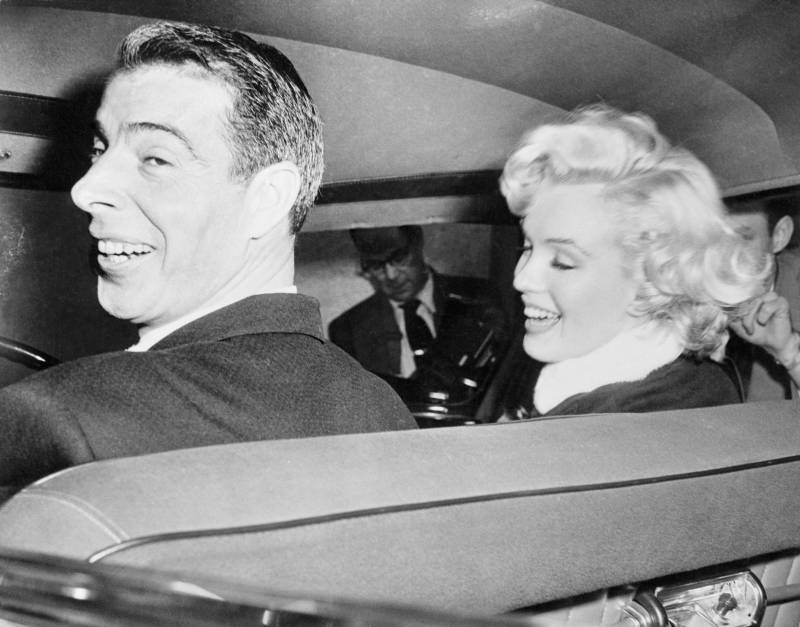 Joe DiMaggio sits in the driver's seat of a Cadillac, Marilyn Monroe smiling by his side. They both look elated. Photographers are visible through the passenger window.