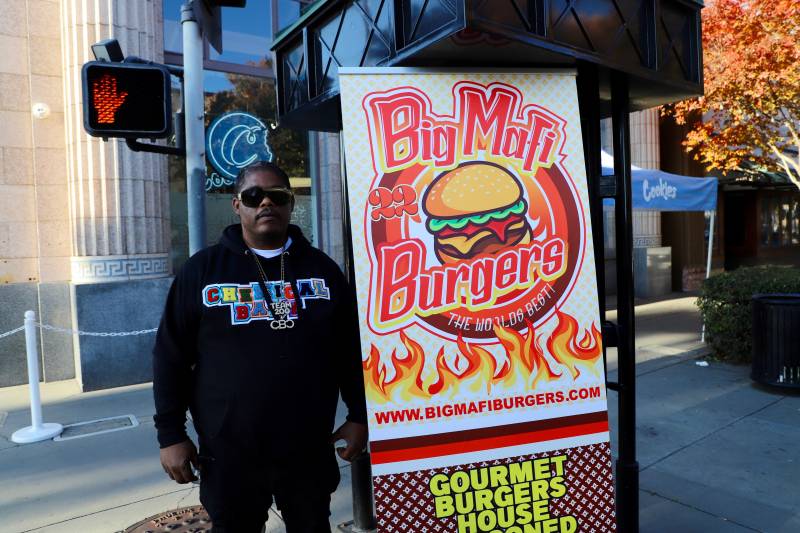 the rapper, Cellski, stands in front of a sign on the street that says "Big Mafi Burgers"