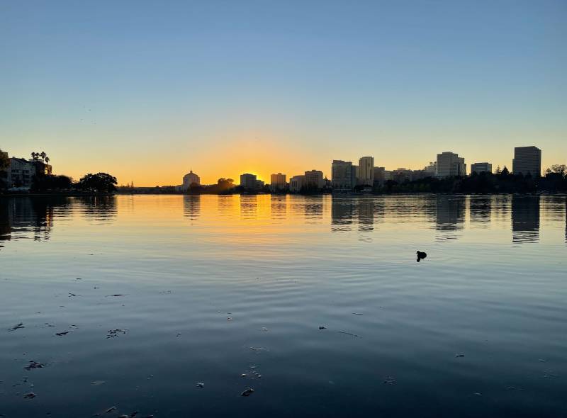The sun sets behind the skyline of Oakland's downtown with Lake Merritt in the foreground