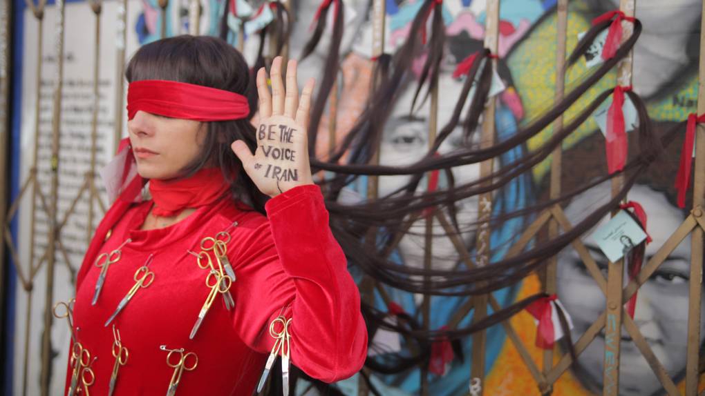Woman in red dress with red blindfold and hair tied to gate holds up hand with writing: 'Be the voice of Iran'