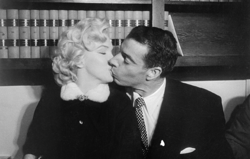 A beautiful blond woman in a high necked suit with soft white collar kisses a man in a formal dark suit with white shirt and dotted tie. Behind them are book shelves holding heavy, formal looking books.