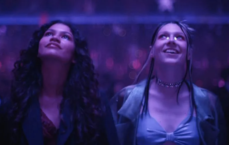 Two young women, bathed in purple light, gaze upwards smiling.