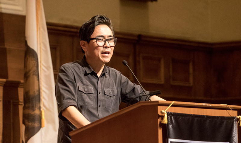 an Asian American man in a gray shirt speaks at a wooden podium