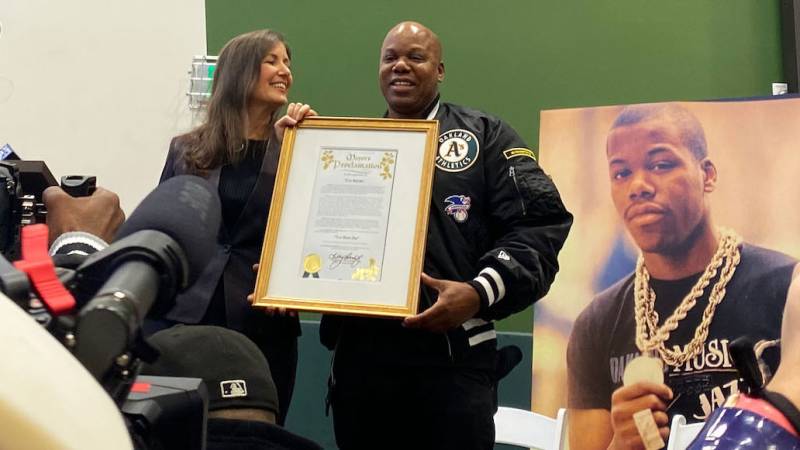 A woman with long hair smiles at a man in an A's starter jacket, holding a framed proclamation from the city