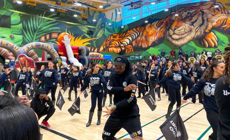 A marching band, dressed in black Too Short shirts, fills a gymnasium with a large Tiger mural in the background