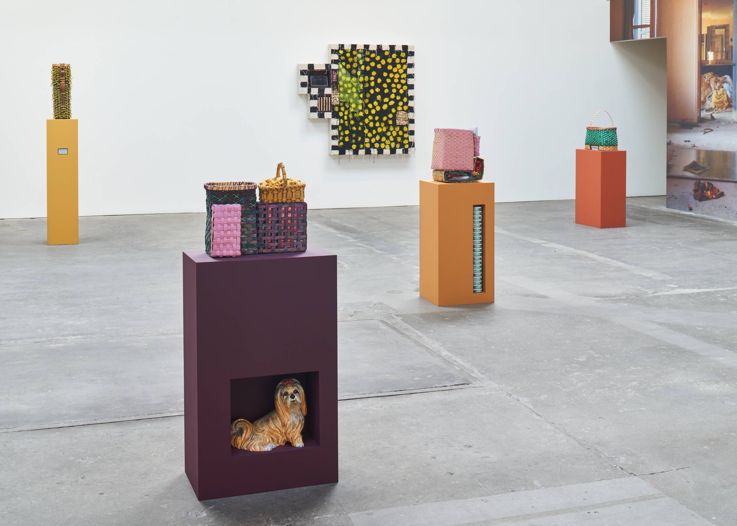 Gallery view of sunset colored pedestals supporting basket sculptures, a textile piece on white wall