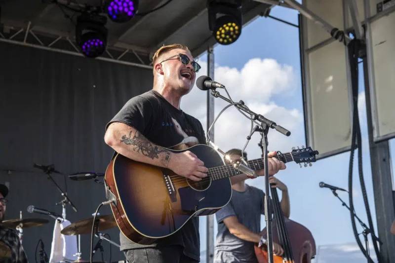 A man in short hair and tattoos plays the acoustic guitar on a festival stage