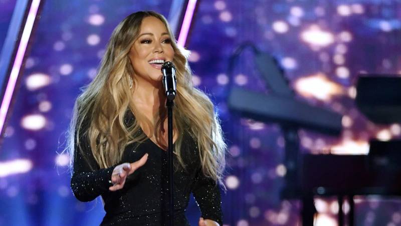 Mariah Carey, a woman in a black sparkly gown, sings into a microphone in front of a purple backdrop