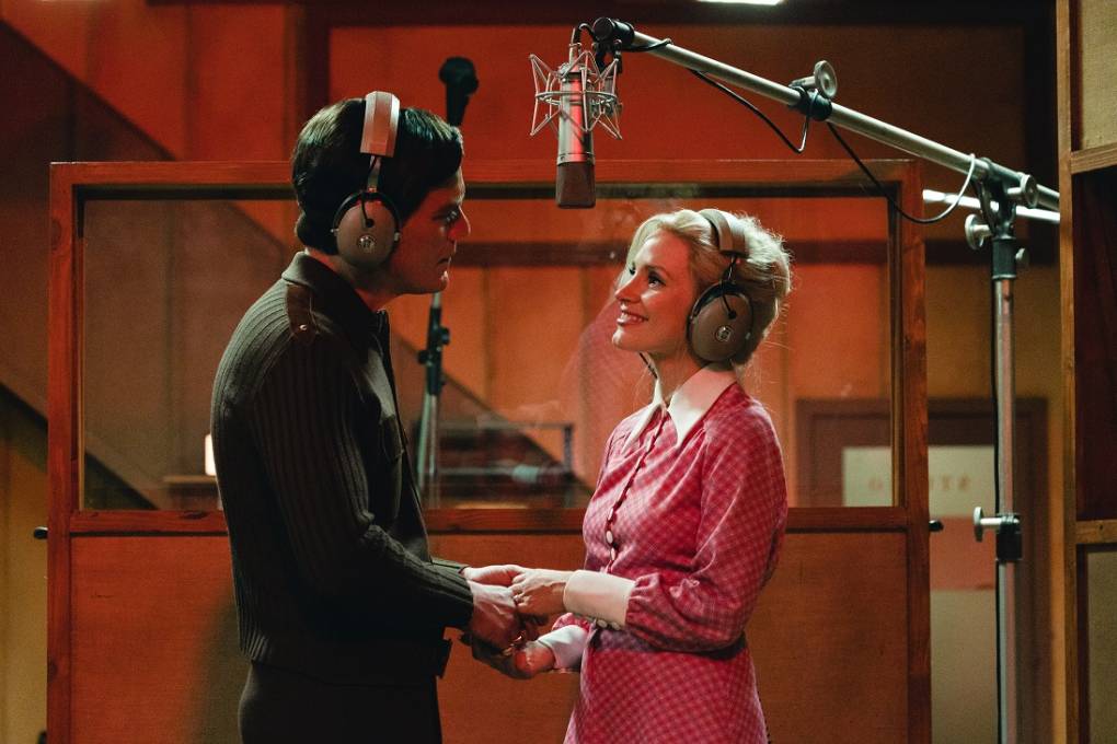 A man with short dark hair, wearing a dark sweater faces a smiling blonde woman in a pink and white dress. They gaze at each other lovingly with a studio microphone hanging over their heads.