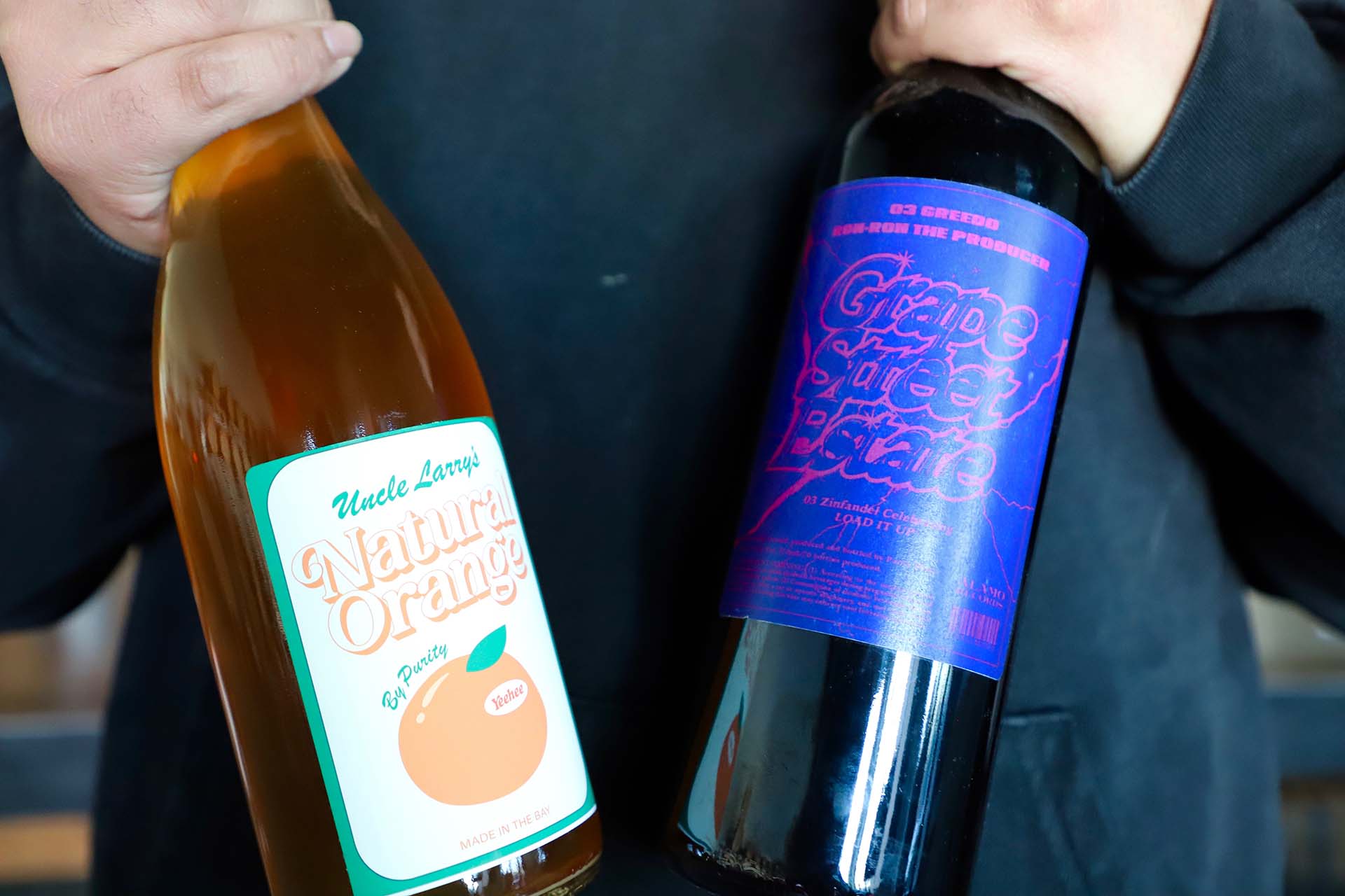 Close-up of two wine bottles; the white and orange label on the left reads "Uncle Larry's Natural Orange" while the purple label on the right reads "Grape Street Estate."
