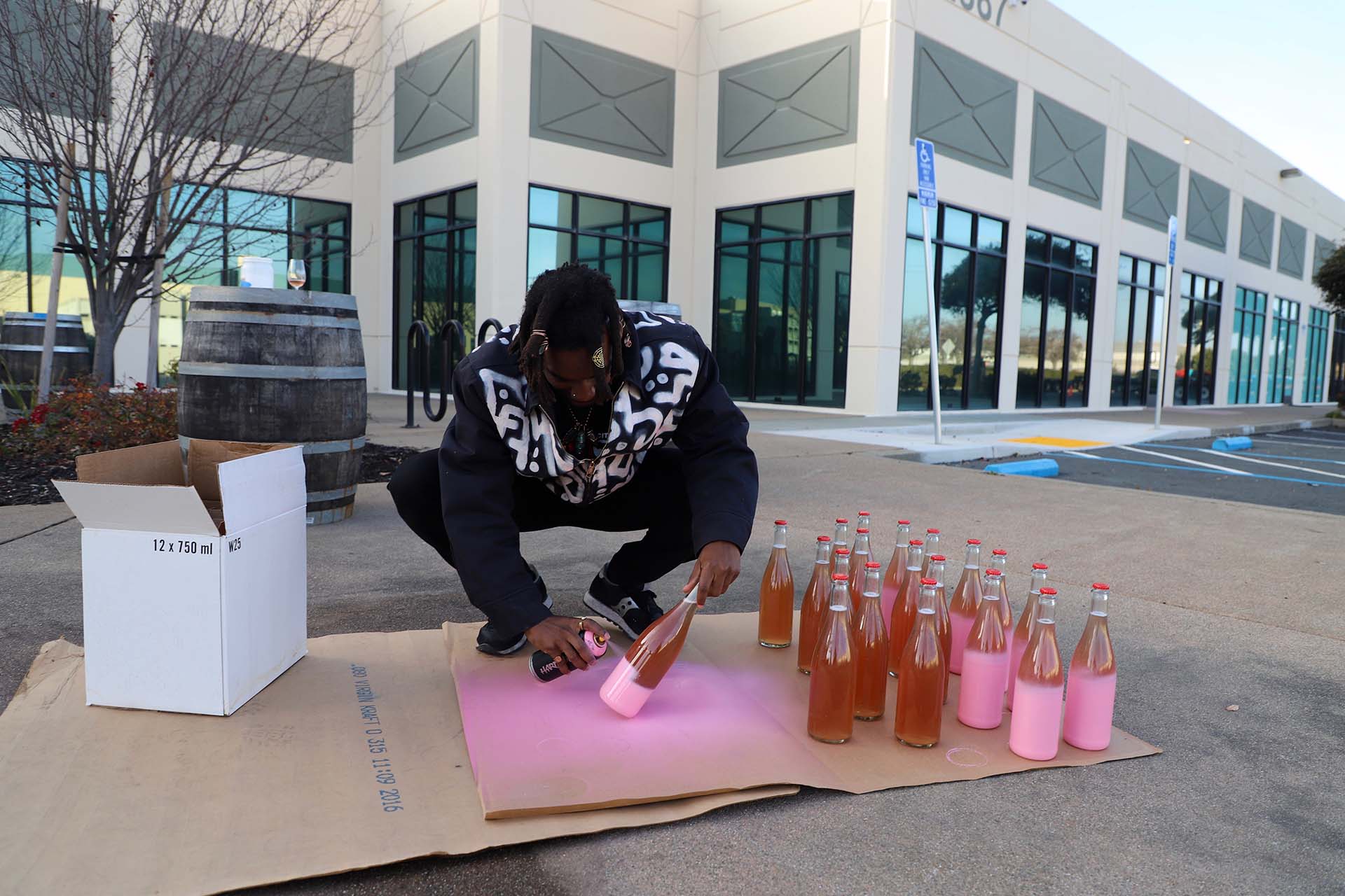 A man crouches down to paint a wine bottle with a can of pink spray paint.