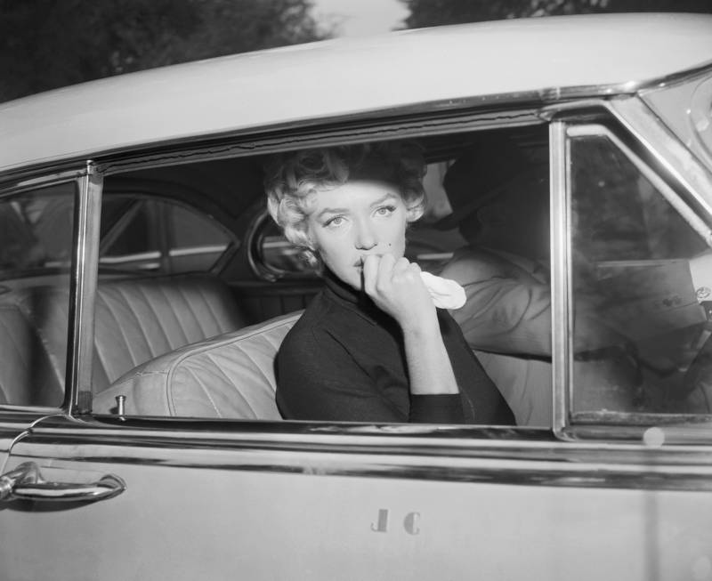 Marilyn Monroe gazes out of a car's passenger side door, white handkerchief held over her mouth, shellshocked expression on her face.