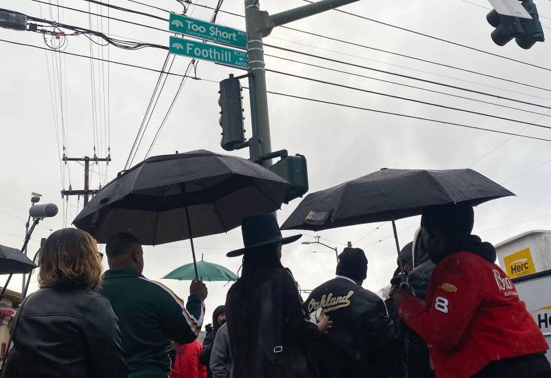 Umbrellas are hoisted below a street sign reading 'Too $hort Way'