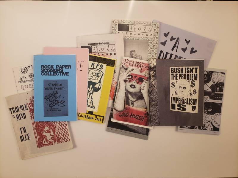 A photograph of 11 fanzines strewn across a table. Their titles include 'Teenage Hussy,' 'Bush Isn't the Problem Imperialism is' and 'Rock Paper Scissors Collective.'