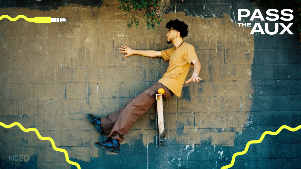 a young Asian-American presenting man in a yellow shirt and brown pants poses against a wall