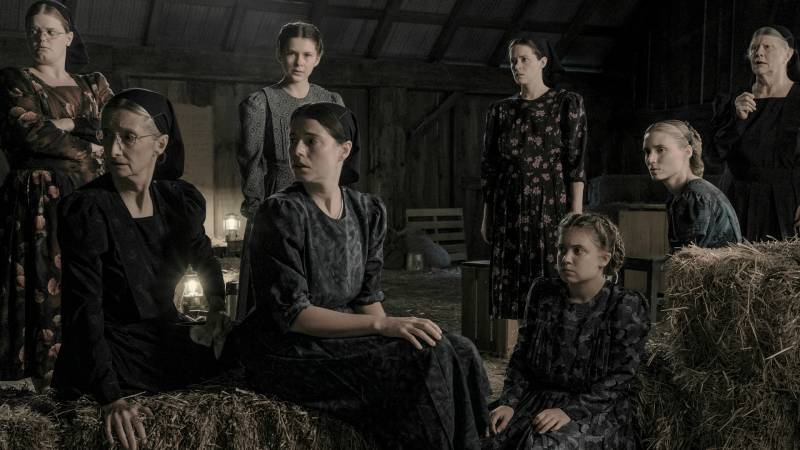 A group of white women of all ages in modest clothing sit in a hay loft, looking left
