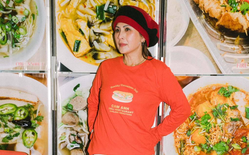 A woman poses in a poppy orange long-sleeved tee, in front of a wall showing pictures of Vietnamese dishes.
