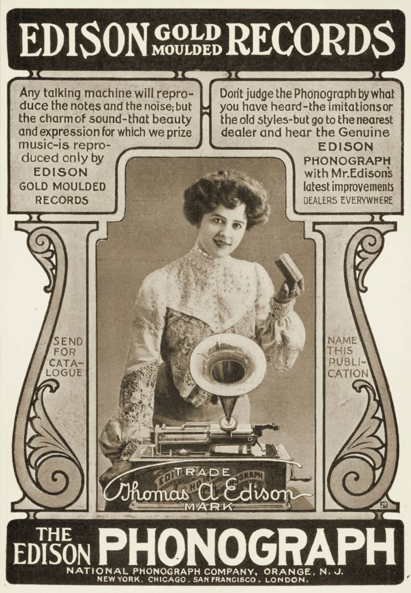 A woman holding an Edison Gold Moulded cylinder record that can be played on the phonograph that stands on a table in front of her.