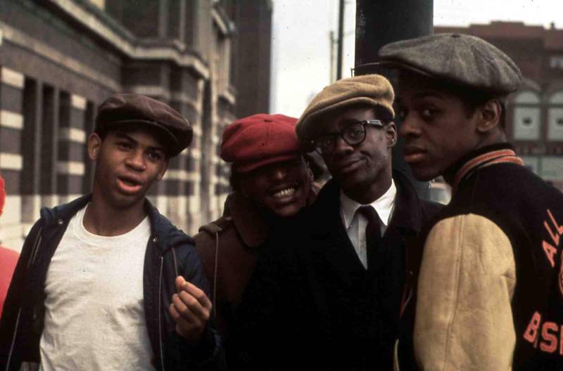 Four young Black people in caps smirk and smile at camera