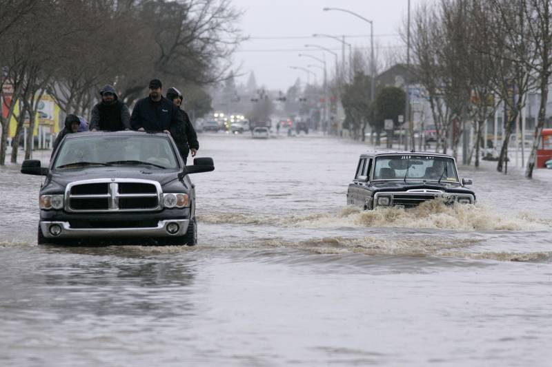 Partially submerged SUVs work their way down a flooded road. Four men stand on the back of one vehicle.