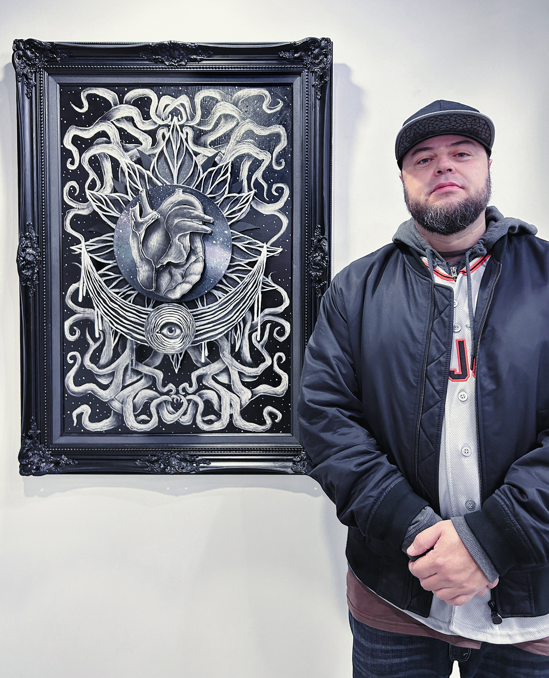 A man with a beard stands beside a framed, black and white ornate drawing