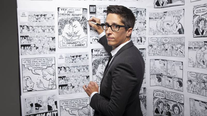 Woman with short hair and glasses in suit in front of wall plastered with comic drawings