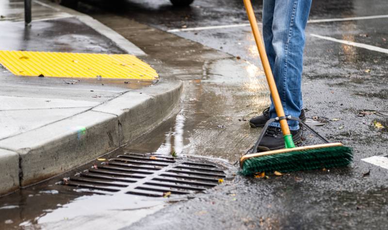 a person in jeans is seen from the waist down, sweeping debris from a wet road near a storm drain