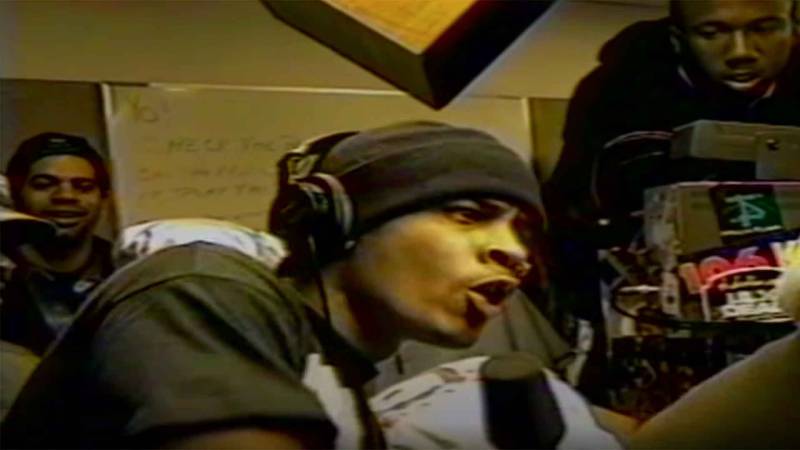 A man in a beanie and headphones raps assertively into a microphone in a radio station studio