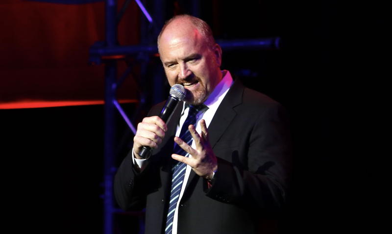 A bald middle-aged man in a suit gestures while speaking into a microphone on a dark stage.