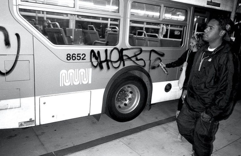 A man stands casually wearing black clothing and an San Francisco 49ers baseball cap, while a woman casually tags a Muni bus in San Francisco. The bus doors are still open.