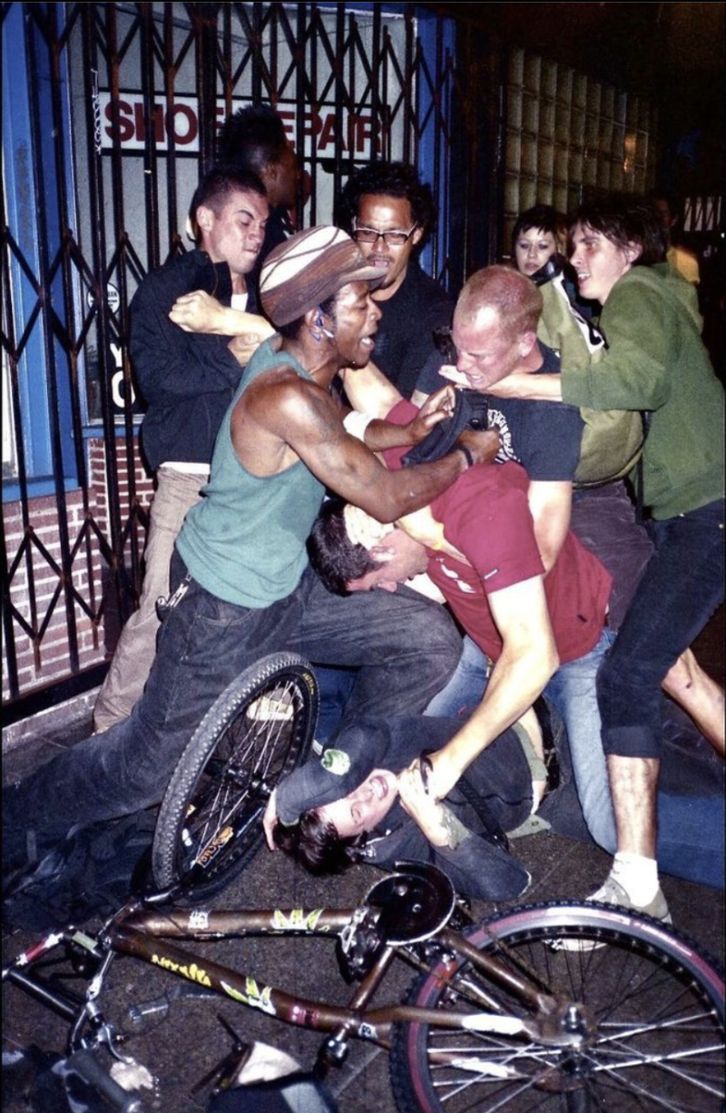 A cluster of seven people push and pull each other in a street fight. A woman lies on the ground underneath it all, a bicycle laying on the ground in front of her.