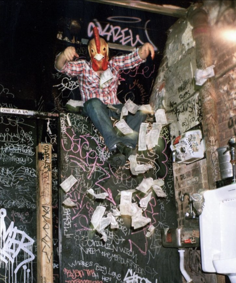 A man wearing blue jeans, flannel shirt and a chicken head mask perches on top of a graffitied bathroom stall, money falling from his hands.