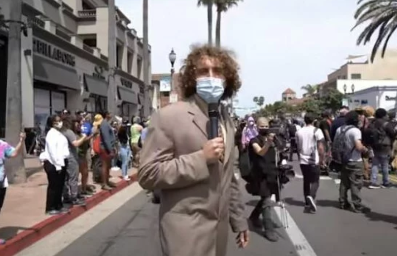 A man in an oversized light brown suit walks through a crowd in the street, wearing a surgical mask and talking into a microphone.